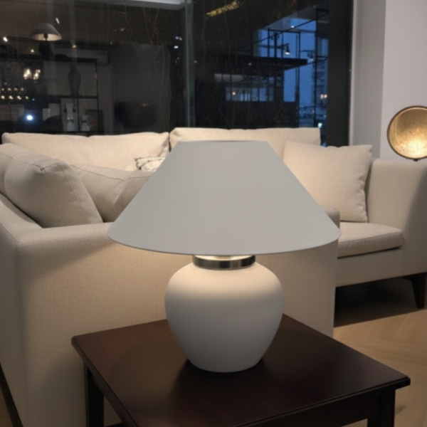 Modern matte white mushroom-style table lamp with a wide tapered shade and chrome neck detail, sitting on a blue round coffee table in a modern living room setting.