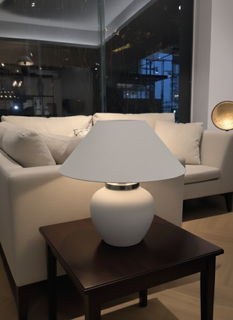 Modern matte white mushroom-style table lamp with a wide tapered shade and chrome neck detail, sitting on a blue round coffee table in a modern living room setting.