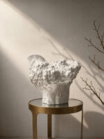 Large white sculptural ceramic vase with an organic coral-textured body and dramatically flared irregular rim, displayed on a brass side table against a warm plaster wall with raking natural light and bare winter branches.