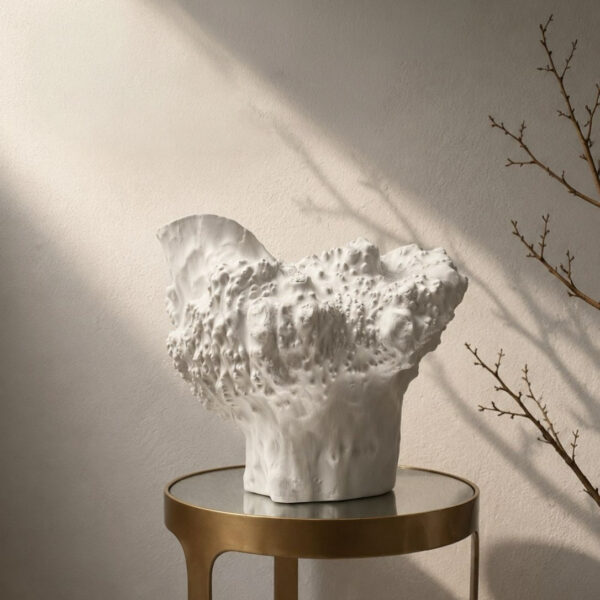 Large white sculptural ceramic vase with an organic coral-textured body and dramatically flared irregular rim, displayed on a brass side table against a warm plaster wall with raking natural light and bare winter branches.