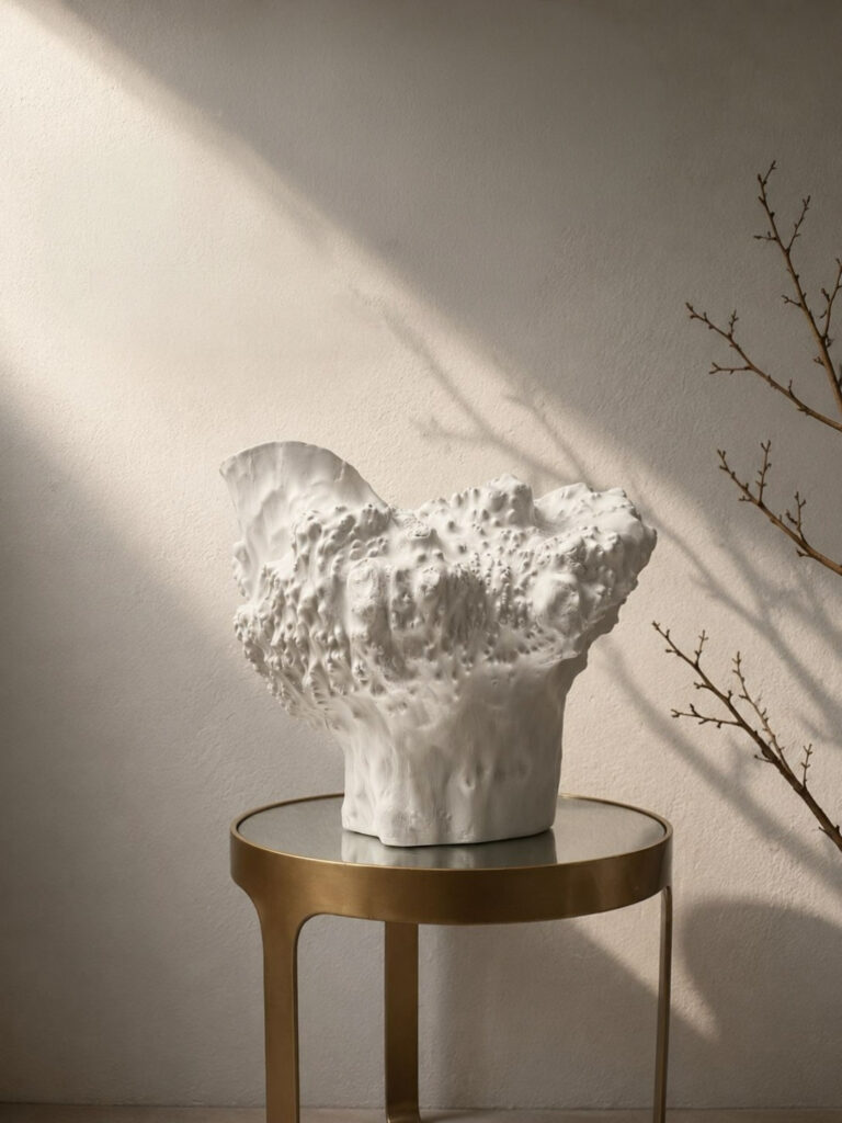 Large white sculptural ceramic vase with an organic coral-textured body and dramatically flared irregular rim, displayed on a brass side table against a warm plaster wall with raking natural light and bare winter branches.