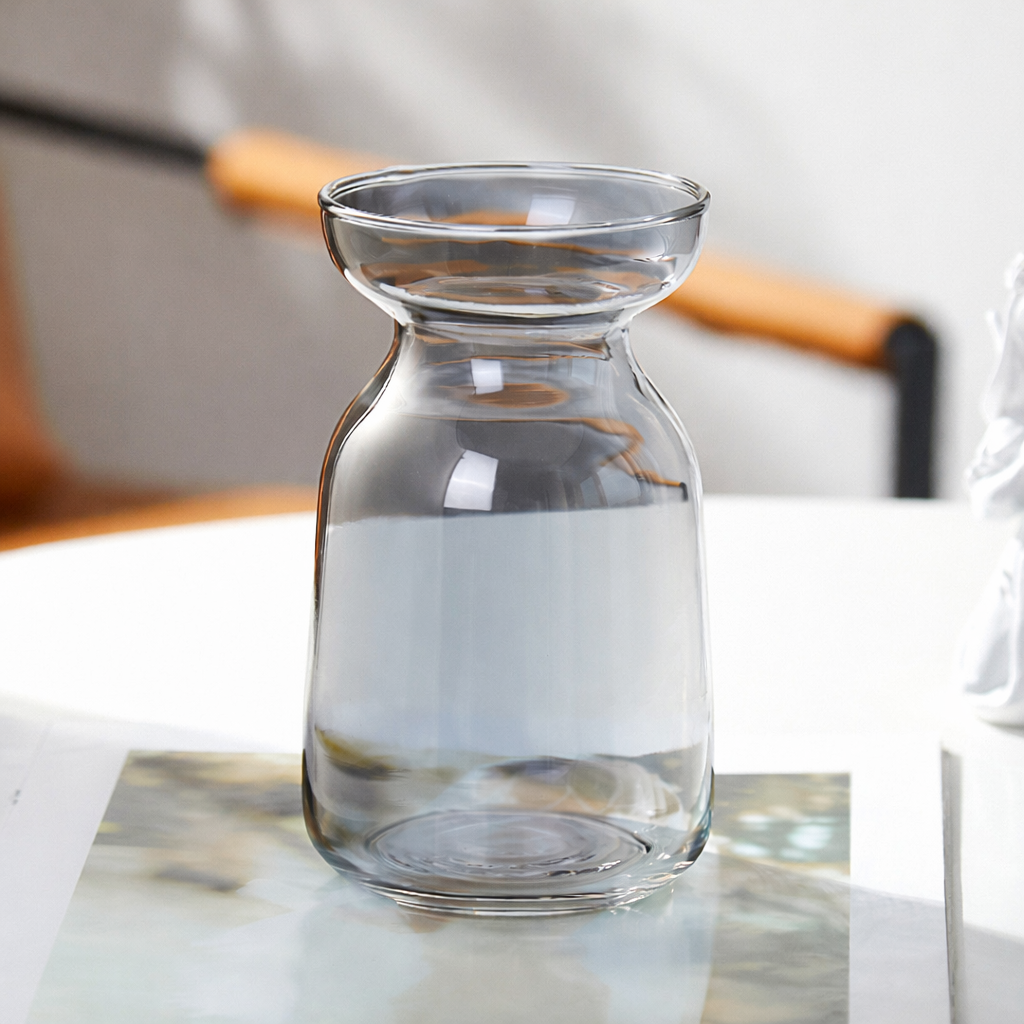 Vase Minimalist clear glass flower vase with a wide flared rim and rounded apothecary-style body, placed on a white marble surface beside an open book in a soft natural light interior setting.