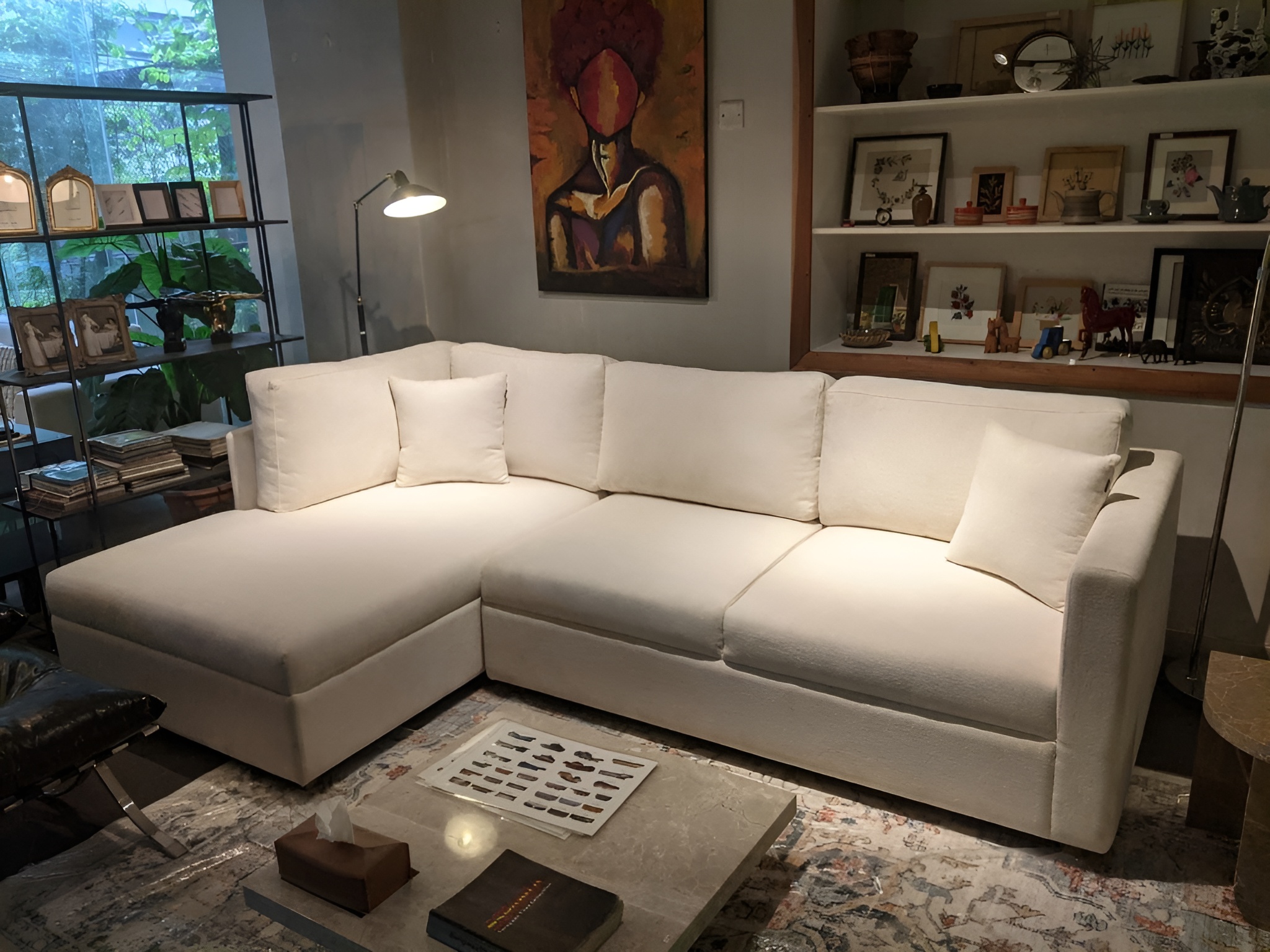WhatsApp Image 2026-01-21 at 2.27.36 PM White boucle L-shaped sectional sofa with a left-facing chaise lounge and plush scatter cushions, styled in an eclectic living space with original artwork, open shelving, and a marble coffee table.