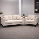 Cream sofa and armchair set with black tapered legs, both pieces upholstered in a soft oatmeal fabric with scatter cushions, styled together in a clean showroom setting with a side table and greenery.