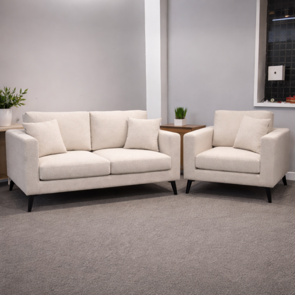Cream sofa and armchair set with black tapered legs, both pieces upholstered in a soft oatmeal fabric with scatter cushions, styled together in a clean showroom setting with a side table and greenery.