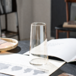 Tapered clear glass vase with a wide open rim narrowing to a rounded base, placed empty on a dark table beside an open book and a woven tray in a moody editorial interior setting.