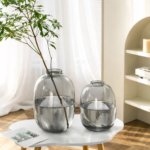 Large smoky grey round glass vase with an organic bubble silhouette and narrow neck, holding a tall leafy branch stem, displayed on a white side table in a bright Scandinavian-style living room.