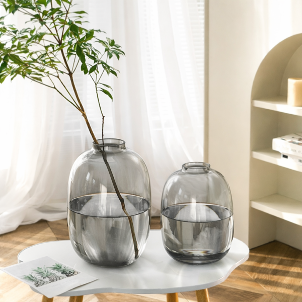 Large smoky grey round glass vase with an organic bubble silhouette and narrow neck, holding a tall leafy branch stem, displayed on a white side table in a bright Scandinavian-style living room.