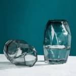 Tall teal geometric faceted glass vase with multi-panel jewel-cut surface in smoky blue-grey, filled with water and standing upright on a white marble surface against a deep teal background.